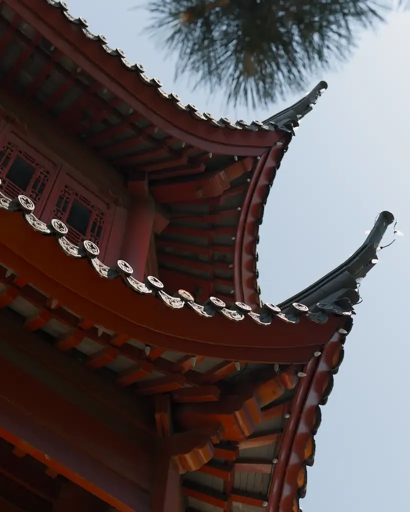 AI generated alt: The upswept corner eaves of a red Chinese pavilion seen from below against a pale sky, tile ends running along the ridge.