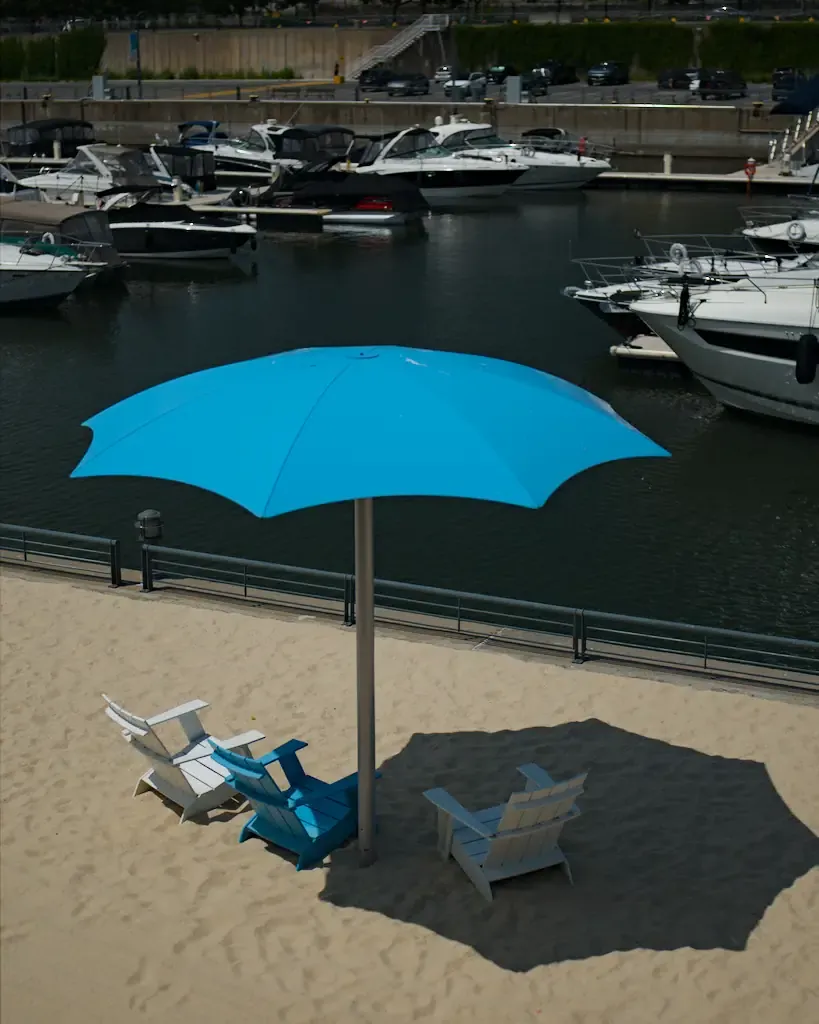 AI generated alt: a bright blue parasol throwing a hard shadow across pale sand, three deck chairs beneath it, moored motorboats on dark water behind.