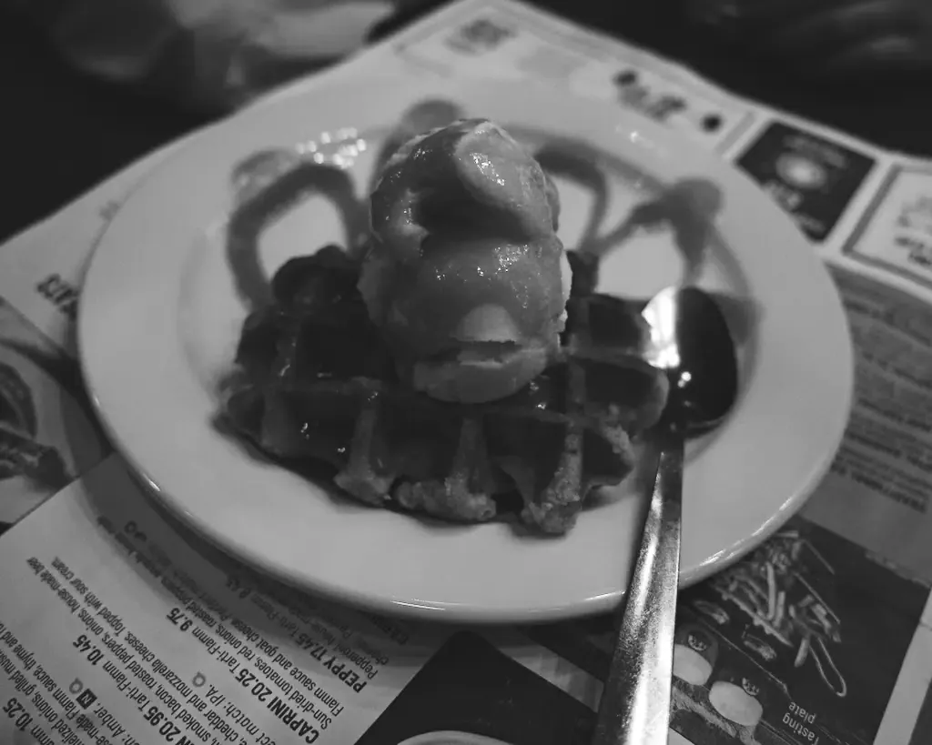 AI generated alt: Black and white: a waffle with a scoop of ice cream and sauce on a white plate, resting on an open printed menu, a spoon across the rim.