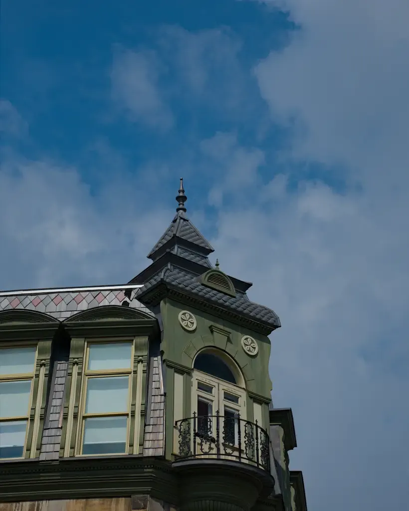 AI generated alt: The green corner turret of an old building against a blue sky with thin cloud, a small wrought-iron balcony below its spire.